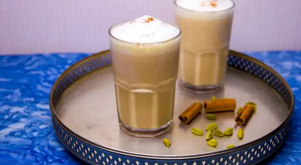 Två glas med krämig chai latte, toppad med skum och kanel. Kardemumma och kanelstänger på brickan ger en hint om detta chai latte recept.