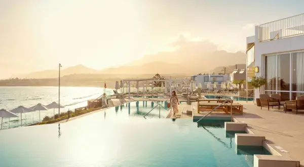 En kvinna i en blommig klänning går mot en infinitypool vid havet på Kreta, Makrigialos. Solnedgången färgar himlen gyllene.