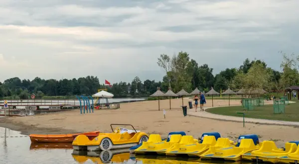 Żółte rowerki wodne i łódki czekają na plaży nad jeziorem blisko Warszawy. Para spaceruje po piasku pod parasolami.