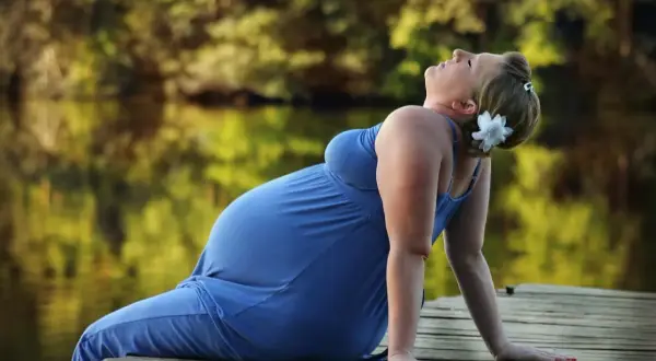 Gravid kvinna i blå klänning på en brygga, med en blomma i håret. Hon lutar sig bakåt, kanske känner hon av Braxton Hicks-sammandragningar.