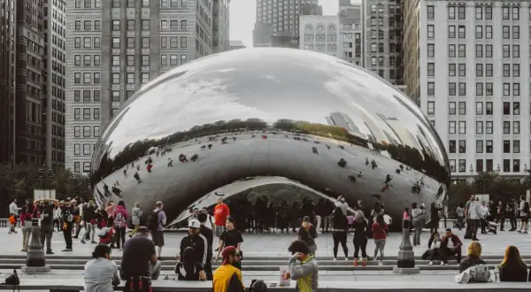 A crowd gathers around Cloud Gate, a reflective public art installation in Chicago, mirroring the city's skyscrapers and sky.