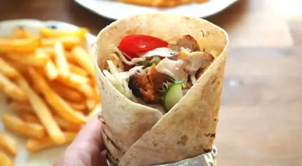 Ręka trzymająca prawdziwy kebab z kurczakiem, warzywami i frytkami w tle.