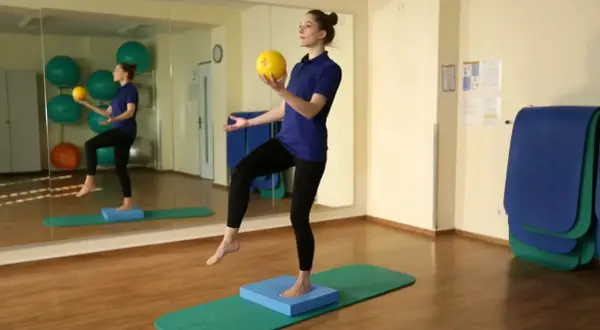 Frau balanciert auf einem blauen Kissen, um zu zeigen, wie einfach arthrose heilen ist. Sie hält einen gelben Ball.