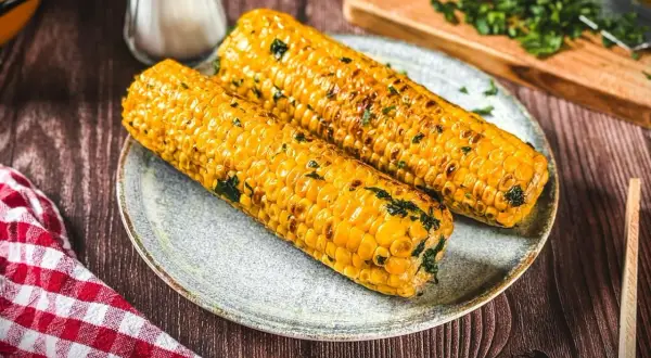 Zwei goldgelbe Maiskolben, perfekt gegrillt und mit Kräutern bestreut, liegen auf einem Teller. Ein Genuss für alle, die Maiskolben grillen lieben!