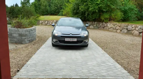 Grå bil parkerad på en vacker betong uppfart. Omgiven av grönskande träd och en stenmur.