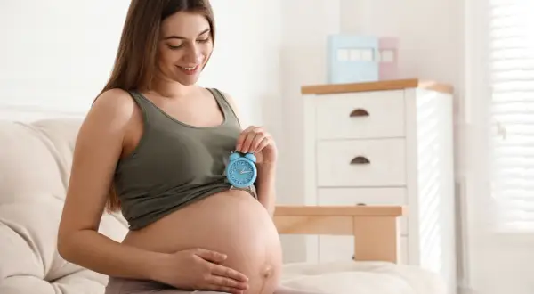 Uśmiechnięta kobieta w 9 miesiącu ciąży trzyma niebieski budzik na brzuchu. Czas na poród!