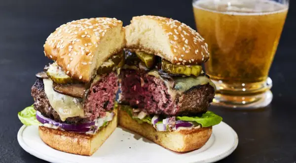 Przekrojony soczysty burger z serem, ogórkiem i grzybami, obok szklanka piwa.