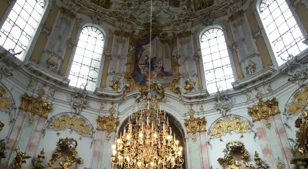 Prunkvoller Kronleuchter unter einer Decke mit Fresken, umgeben von opulenten Verzierungen, die die **Rokoko Architektur** widerspiegeln.