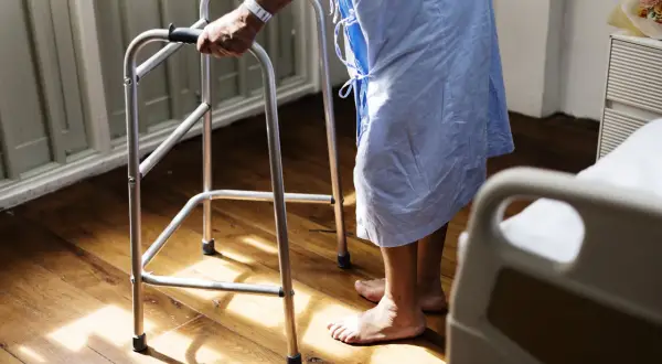 Osoba w szpitalnej koszuli, wspierając się na chodziku, stawia pierwsze kroki po udarze.