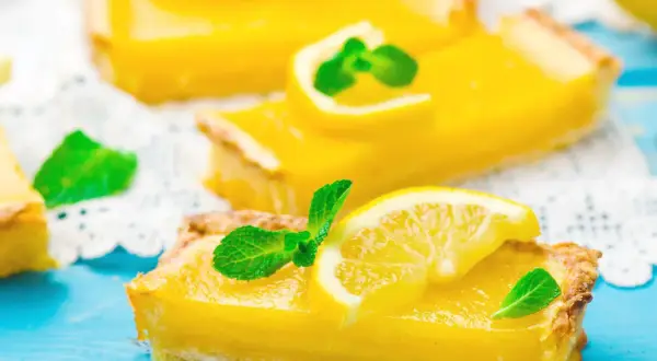 Fette di torta fredda al limone, guarnite con fettine di limone e menta fresca, su un tagliere blu.