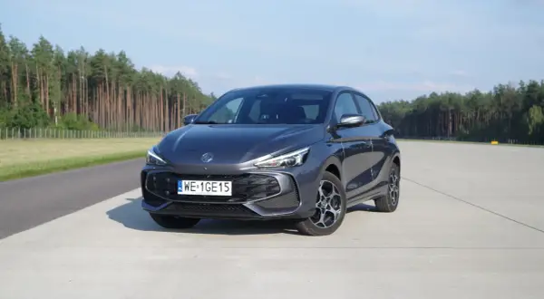 Szary MG3 Hybrid na betonowym placu, z lasem w tle.
