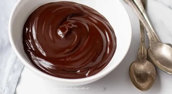 Una ciotola bianca piena di glassa al cioccolato veloce, lucida e invitante, con due cucchiai d'argento accanto.