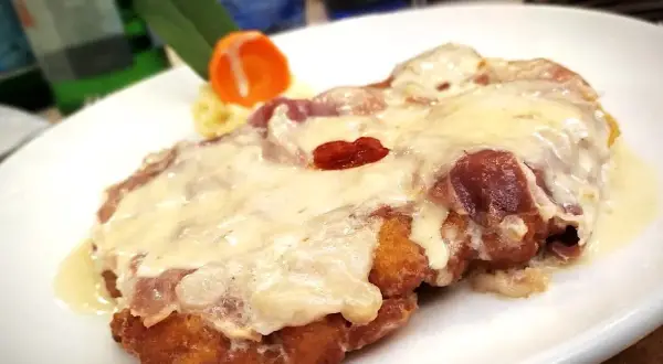 Cotoletta alla bolognese dorata e filante, con prosciutto e formaggio fuso, servita su un piatto bianco.