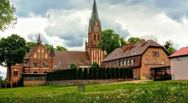 Majestatyczne sanktuarium w Gietrzwałdzie, z ceglaną architekturą i strzelistą wieżą, góruje nad zieloną łąką pełną kwiatów.