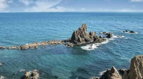 Formaciones rocosas emergen del mar turquesa en la **sierra de Cabo de Gata**. El sol ilumina las aguas cristalinas y el cielo azul con nubes blancas.