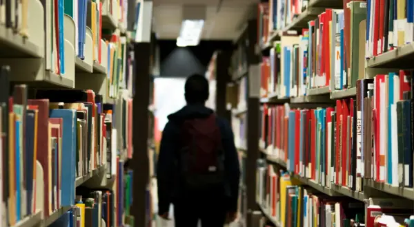 Ein Schüler mit Rucksack geht durch eine Bibliothek. Vielleicht sucht er nach Büchern, die ihm zeigen, dass das Gymnasium viel schwerer als Realschule ist.