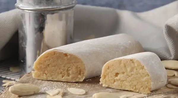 Pâte d'amande prête à être utilisée pour recouvrir un gâteau. Des amandes effilées et un tamis à sucre complètent cette scène gourmande.