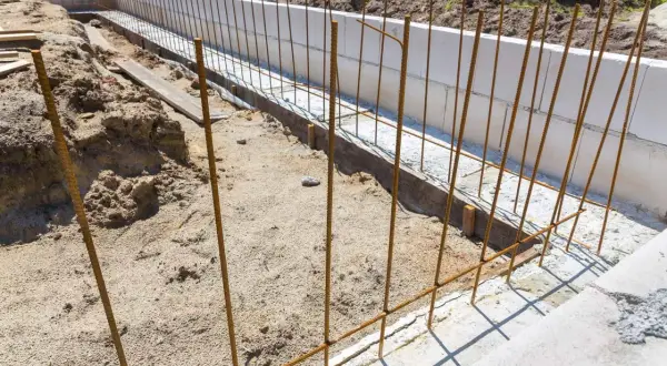 Budowa fundamentów: pionowe pręty zbrojeniowe czekają na beton. Widać też białe bloczki fundamentowe i piasek.