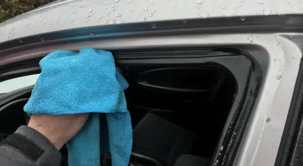 Ręka z niebieską ściereczką czyści uszczelki drzwi samochodu. Dowiedz się, czym smarować uszczelki w samochodzie domowe sposoby, by zapobiec skrzypieniu.