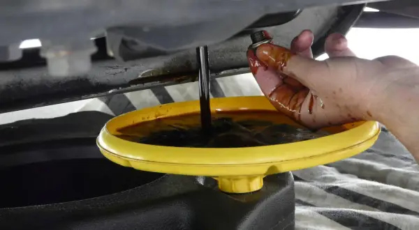 Mano sporca d'olio che tiene il tappo di scarico mentre l'olio scuro cola in una bacinella gialla. Forse c'era troppo olio nel motore.