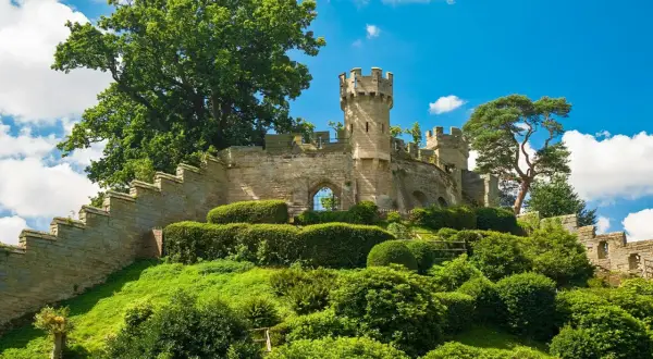 Warwick Castle reser sig stolt på en grönskande kulle, omgiven av frodig grönska och en klarblå himmel med vita moln.