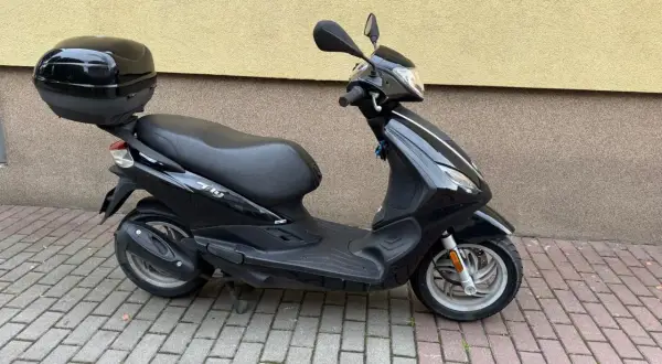 Czarny skuter Piaggio Fly 50 z kuferkiem na bagaż, zaparkowany na chodniku z kostki brukowej.