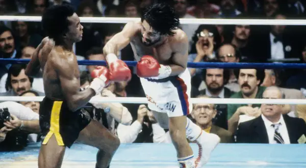 Sugar Ray Leonard lands a punch on Roberto Duran in a fierce boxing match. The crowd watches intently as the two legends battle in the ring.