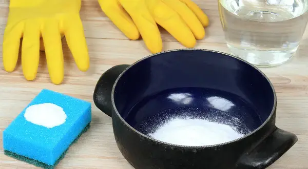 Gants jaunes, éponge bleue avec bicarbonate, casserole noire et un bocal de liquide. Astuces pour savoir comment nettoyer une casserole qui a brûlé.