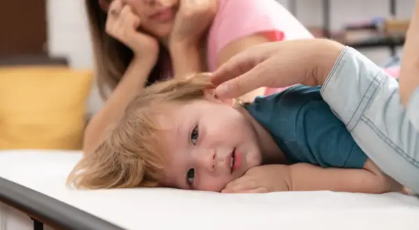 Małe dziecko z blond włosami leży na białym materacu, jego mama patrzy z troską. Czy to początek pandas choroba?