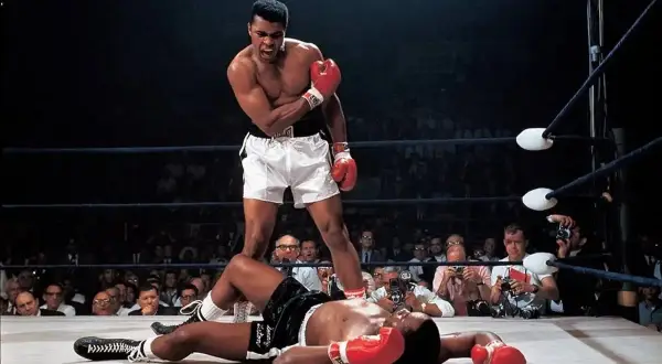 Iconic Muhammad Ali stands over a fallen opponent in the ring, a triumphant roar escaping his lips. A crowd watches, cameras flashing.