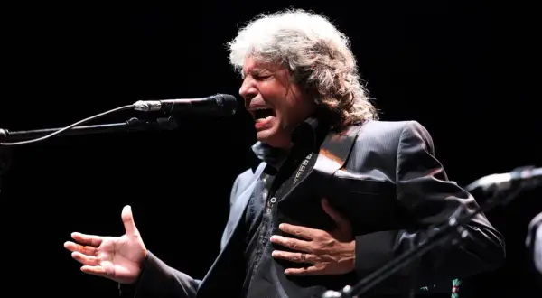 Un cantante de grupos flamencos de los 80, con melena rubia, canta apasionadamente frente a un micrófono, con la mano en el pecho.