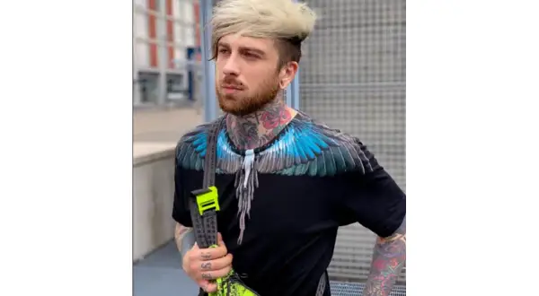 Hombre con tatuajes visibles en cuello y brazos, viste camiseta negra con alas azules y bolso neón.
