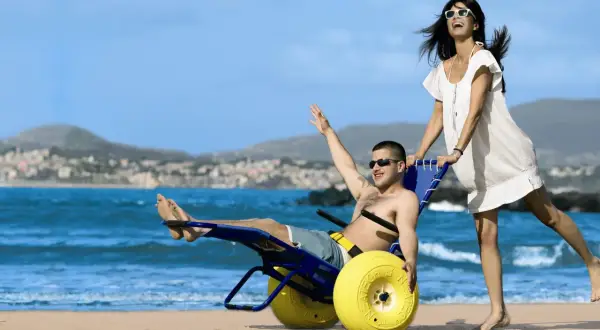 Coppia felice sulla spiaggia, lui su una sedia job con ruote gialle, lei lo spinge. Mare e collina sullo sfondo.