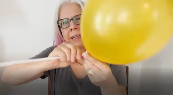 Kobieta w okularach mocuje żółty balon foliowy do białego patyka.