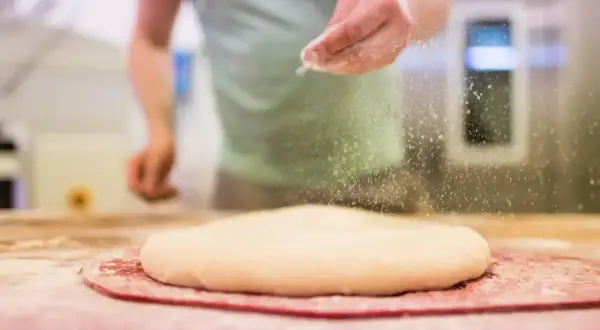 Ręka posypuje ciasto na pizzę mąką. Odpowiednia jaka mąka do pizzy to klucz do sukcesu.