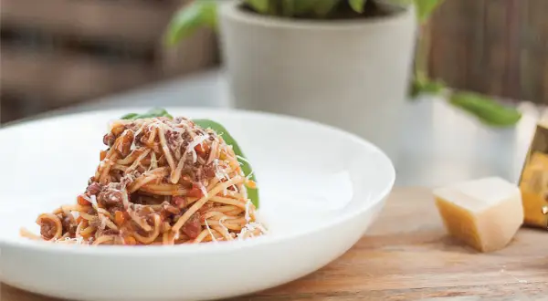 Pyszne spaghetti bolognese przepis oryginalny, podane na białym talerzu, posypane parmezanem. Obok kawałek sera i bazylia.