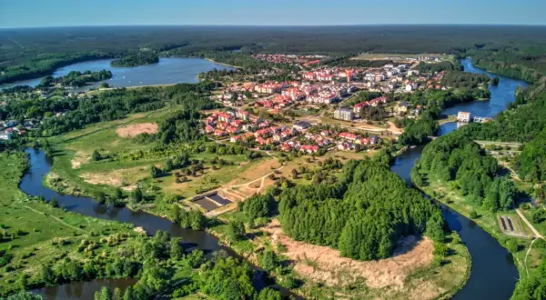 Malownicze miasteczko z czerwonymi dachami rozciąga się nad krętą rzeką, otoczone zielonymi lasami i polami.