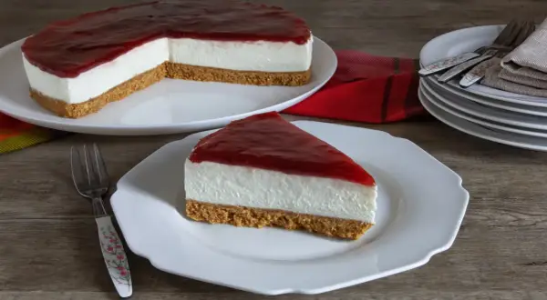 Una fetta di cheesecake fredda con copertura rossa su un piatto bianco, accanto a una torta intera.