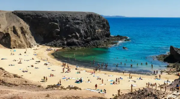 Plaża z turystami kąpiącymi się w turkusowej wodzie, otoczona skalistymi klifami.