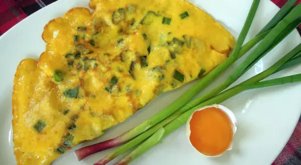 Une omelette à l'aillet, garnie de ciboule verte, est servie sur une assiette blanche avec des tiges d'aillet et un jaune d'œuf.