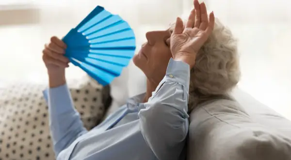 Starsza kobieta zmagająca się z uderzeniami gorąca, wachlująca się niebieskim wachlarzem. Może to być związane z menopauzą, którą leczy fezolinetant.