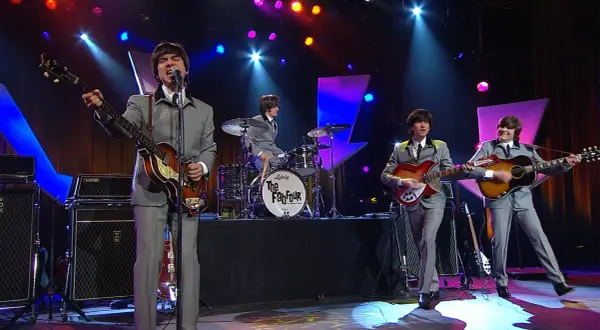 Una cover band sul palco, con abiti grigi e chitarre, ricorda i Fab Four.