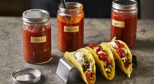 Tre burkar med tacosås, märkta "Mild", "Medium" och "Hot", står bredvid färdiga tacos. Perfekt för att hitta ditt tacosås recept.