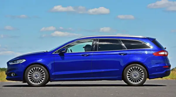 Niebieski kombi Ford Mondeo na tle błękitnego nieba z chmurami. Kierowca w czapce z daszkiem za kierownicą.