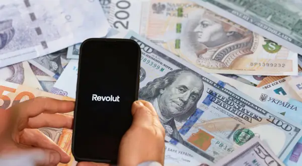 Dłoń trzyma smartfon z logo Revolut na tle różnych walut. To banki, które nie współpracują z komornikiem, oferując alternatywę dla tradycyjnych instytucji.
