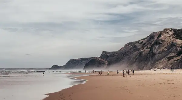 Plaża w Faro, wrzesień. Ludzie grają w piłkę, spacerują po piasku. W tle skaliste klify i ocean.