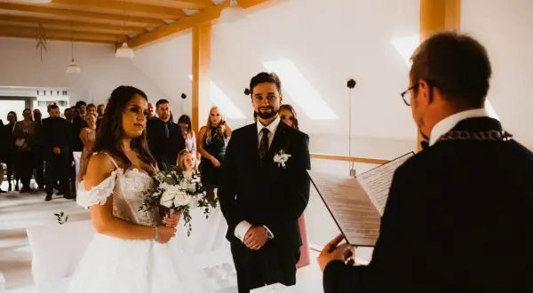 Para młoda podczas ceremonii cywilnej. Panna młoda w białej sukni, pan młody w garniturze. Goście obserwują. Czy po ślubie cywilnym można być chrzestnym?