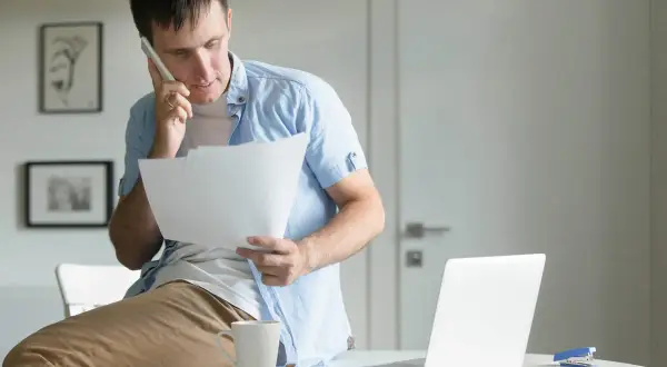 Uomo al telefono con fogli in mano, preoccupato per una busta paga errata in eccesso.