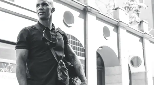 Nico Musoke, en man med tatueringar, bär en väska framför en byggnad i svartvitt.