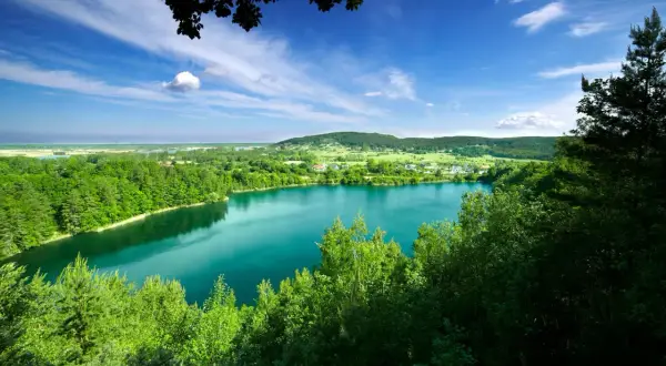 Turkusowe jezioro w Międzyzdrojach otoczone zielenią, z widokiem na wzgórza i błękitne niebo z chmurami.